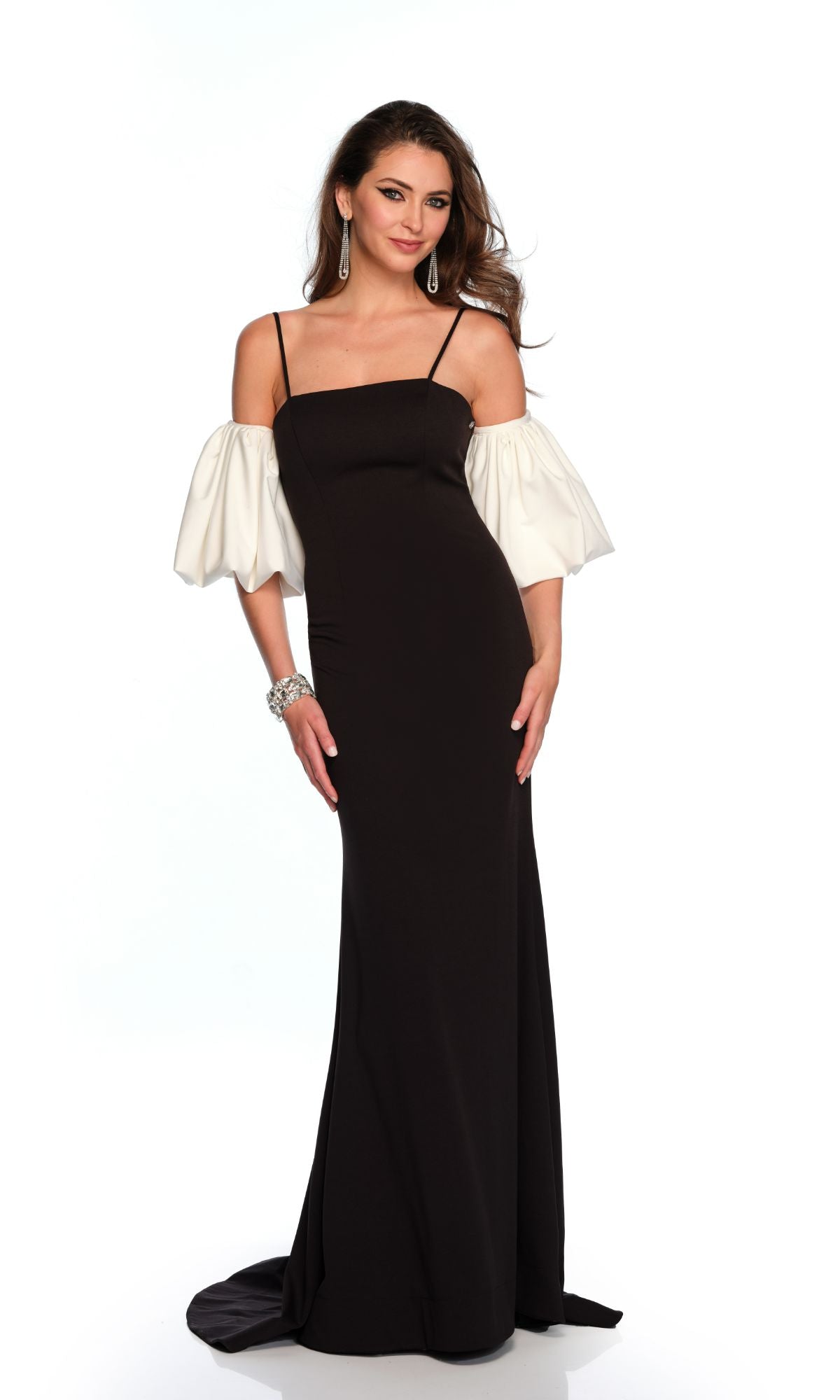 A woman wearing the Long Formal Dress 11368 by Dave and Johnny, a black fitted gown with white off-the-shoulder puff sleeves and thin straps, paired with statement jewelry, stands against a plain white background.